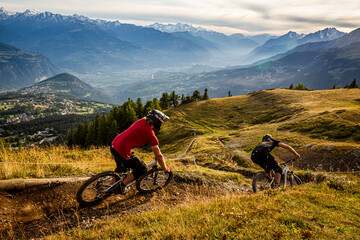 CRANS MONTANA, SWITZERLAND. Two mountain bikers riding a twisty trail in the purpose built bike park. Snow capped mountains and evening mist in the Sion valley behind.