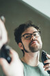 © Paul - a handsome young man with a thick beard and glasses is illuminated on one side by a soft light. fashionable beard, happy. reflection in the mirror. irritation after shaving.  very serious