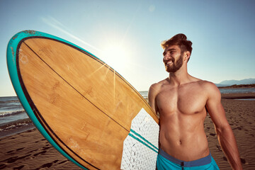 Wall Mural - young caucasian beardy male walking on the sunny beach, holding surfboard, smiling. ultraviolet radiation concept