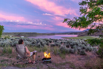 a man sitting by a burning fire watching a scene with sage brush, a tree, a lake and mountains while