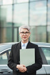© AnnaStills - Portrait of mature businesswoman in suit and in eyeglasses holding documents and looking at camera while standing in the city
