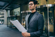 © BullRun - Confident young financial manager holding paper documentation in hands and reading information walking on megalopolis street.Serious intelligent young entrepreneur checking business reports