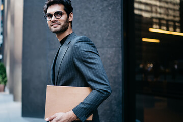 Half length portrait of successful young entrepreneur in elegant suit smiling at camera while holding file with information data.Cheerful proud CEO with folder in hand strolling near office building