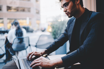 Concentrated male entrepreneur in trendy wear and spectacles typing documentation for software, prosperous owner of business working remotely on netbook using wifi connection sitting in cafe shop