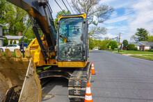 Front View Of A Yellow Heavy Equipment Excavator Parked On The Side Of A Residential Street In Front Of A House With Orange Warning Cones In Front
