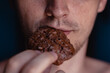 © Kate - A detailed close-up shot of the mouth of a young handsome man who bites a cookie, holding it in his hand on a home background. Man eating chocolate cookie with peanut, white teeth, close up face.