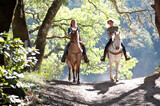 Two women riding horses toward camera with backlit leaves