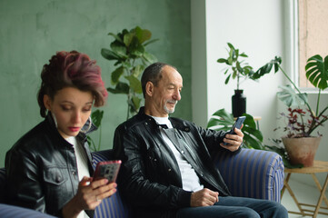 Family portrait of adult daughter and senior father in loft room with houseplants. Man and girl are wearing black leather jackets in punk style and browsing on their smartphones without communication