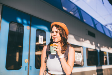 Tourism and travel in the summer. Vacations for the student. Work and travel. Caucasian young woman drinks coffee on the platform of the railway station against the background of the train