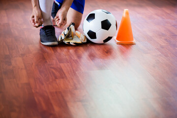 Sticker - Soccer ball with futsal player sitting and tying sport shoelaces in stadium hall. Futsal player and training equipment on the wooden floor.