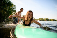 Laughing Woman Lies On Surfboard And Rows Her Hands And Male Instructor Supports Her