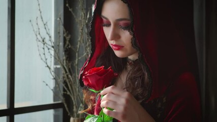 Wall Mural - Beautiful young sexy charming woman, red velvet vintage hood stands near window gothic dark room. Backdrop branches. Girl gently touches rose petals flower. Face closeup, bright evening holiday makeup