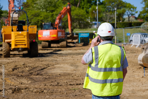 Supervisor wearing safety helmet and hi-vis vest makes mobile phone call on construction site