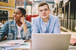 © BullRun - Portrait of handsome young caucasian male student in eyewear sitting next to laughing african american woman doing homework together, hipster guy looking at camera during working process with partner.