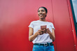 © BullRun - Smiling young African American woman in t-shirt holding mobile phone and looking away, cheerful dark skinned female blogger using smartphone stand on red wall background. Online Internet connection
