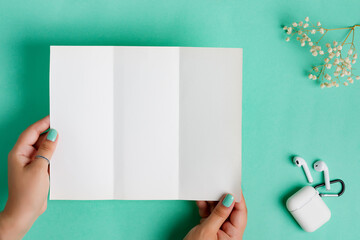 Wall Mural - Empty mock-up paper sheet for letter or message in woman's hands above pastel turquoise background with earphones and dry twig.