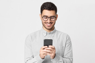 Young man wearing gray shirt and glasses, holding his smart phone and exchanging messages with friends, isolated on studio background