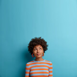 Fototapeta  - Vertical shot of cute dark skinned woman with Afro hairstyle, looks above with pondering expression, notices something interesting, blank space on blue background. Face expressions and emotions