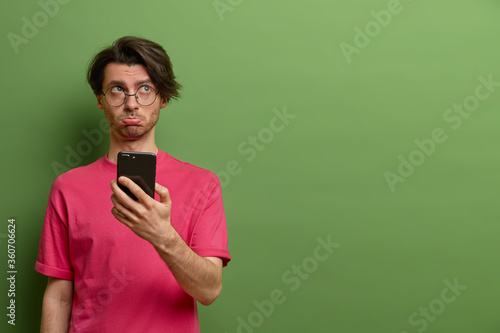 Upset dissatisfied European man holds smartphone with sad expession, finds out someone spreading rumors about him in internet, regrets missing chance to buy tickets online, dressed in pink t shirt