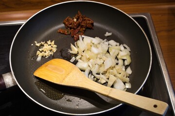 Wall Mural - Closeup shot of oil, onion, garlic and dried tomatoes in a frying pan  with a wooden spoon