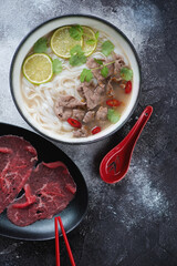 Vietnamese pho bo soup served with slices of raw beef meat, above view on black and white stone background, vertical shot