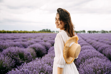 Young brunette woman standing in lavender field, wearing a bohemian white dress and a straw hat.