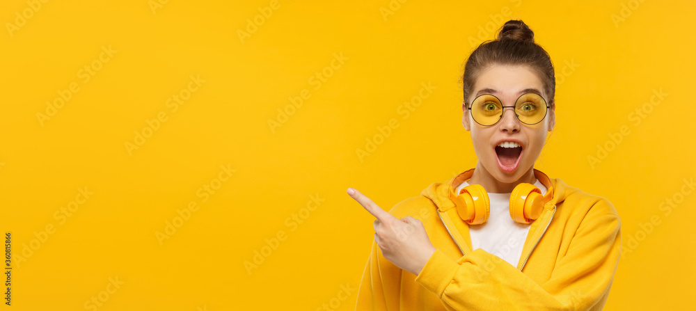 Horizontal banner of young shocked girl, wearing round glasses and headphones on neck, pointing left to copy space, isolated on yellow background - obrazy, fototapety, plakaty 