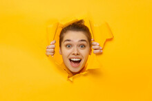 Young Teen Girl Tearing Paper With Hands And Looking Through It With Amazed Face And Round Eyes, Isolated On Yellow Background