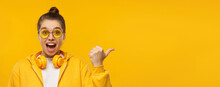Horizontal Banner Of Young Excited Girl Wearing Hoodie, Glasses And Wireless Headphones Around Neck, Pointing Right To Copy Space, Isolated On Yellow Background