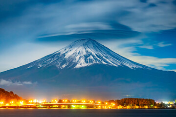 Wall Mural - Japan. Snow on top of Mount Fuji. City Lights Fujikawaguchiko on the background of the volcano. Night bridge on the background of the volcano Fujiyama. Mount Fujiyama close-up. Kawaguchiko Lake