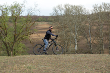 Sports brutal bearded guy on a modern mountain bike. Cyclist on the green hills in the spring.