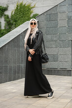 Modern Stylish Muslim Woman In Hijab, Leather Jacket And Black Abaya Walking In City Street , Wearing Sunglasses And Bag, Fashion Style Trend