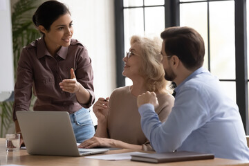 Diverse colleagues with mature businesswoman team leader wearing glasses working on project together, sitting at office desk with laptop, discussing strategy, sharing business ideas, brainstorming