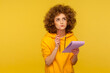 © khosrork - Portrait of pensive curly-haired hipster woman in urban style hoodie thinking over smart idea, holding pencil and notebook to write plans, to-do list. indoor studio shot isolated on yellow background