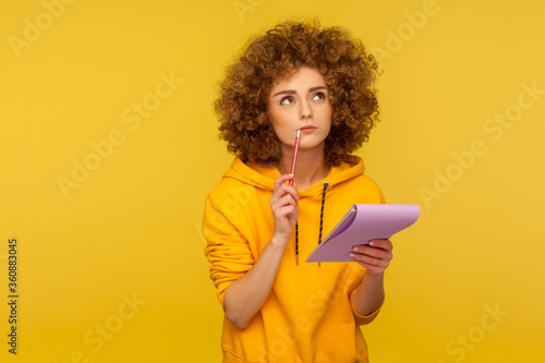 Fotomural Portrait of pensive curly-haired hipster woman in urban style hoodie thinking over smart idea, holding pencil and notebook to write plans, to-do list