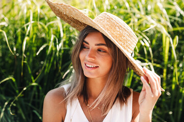 Wall Mural - Young smiling woman wearing straw hat while sitting in park in front of a grass