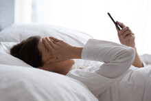 Upset young woman lying in bed feel sick or distressed using modern cellphone gadget, sad ill millennial female relax in bedroom hold smartphone read bad news, receive negative text message