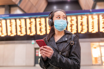 Wall Mural - Chinese woman wearing face mask at train station and maintaining social distance - young asian woman using smartphone and looking away with departure arrivals board behind - health and travel concepts