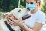 Fototapeta Zwierzęta - Close up photo of a caucasian blonde woman with medical mask on face playing outside with her dog