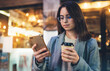 © A_B_C - Young girl drink coffee outdoors using mobile smartphone on background bokeh light in night city, woman hold in hands sellphone, online wi-fi internet
