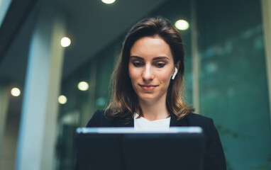 Successful female banker using tablet and wireless earphones outdoors near his office background lights, portrait young woman professional manager working on pc computer evening city