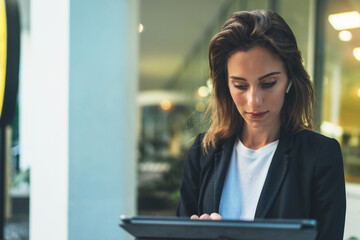 Wall Mural - Lifestyle business woman using tablet walking in evening city, girl banker dressed in business style listening to music wireless earphones and typing text message on laptop device, flare light