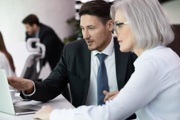 Business man and woman colleagues sitting at desk discuss startup using laptop. Businessman sharing ideas with mature female coworker. Two serious businesspeople solving issues working on project