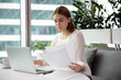 © fizkes - Focused millennial one female worker busy at laptop in modern office room. Woman manager or ceo sit and hold paper sheet or document use computer business networking. Businesswoman at desk read report