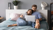 © fizkes - Young man and woman hugging, relaxing, lying in cozy bed in bedroom, using laptop, looking at screen, young couple watching movie, shopping online, enjoying leisure time, lazy weekend at home