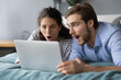 Shocked young couple looking at laptop screen, lying on bed at home, surprised woman and man with open mouths reading unexpected news, online lottery win, amazed by unbelievable shopping offer