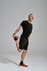 Full length shot of middle aged muscular man in black sportswear looking aside while stretching his body before workout in studio over grey background