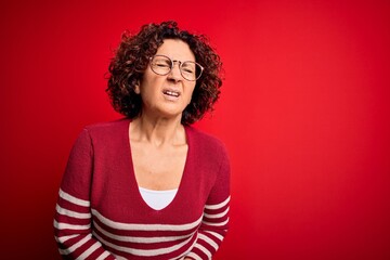 Poster - Middle age beautiful curly hair woman wearing casual striped sweater over red background with hand on stomach because nausea, painful disease feeling unwell. Ache concept.