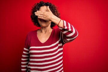 Wall Mural - Middle age beautiful curly hair woman wearing casual striped sweater over red background smiling and laughing with hand on face covering eyes for surprise. Blind concept.