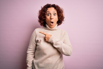 Canvas Print - Middle age beautiful curly hair woman wearing casual turtleneck sweater over pink background Surprised pointing with finger to the side, open mouth amazed expression.